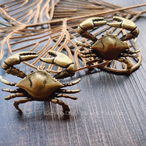 Peut inclure: Deux figurines de crabes en bronze avec des pinces et des corps détaillés sont présentées sur une surface en bois. Les crabes sont placés près d'une feuille décorative de couleur or. L'esthétique générale est rustique et nautique.