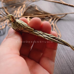 May include: A detailed, antique-style brass bird figurine with long tail feathers. The bird is held in a hand, showcasing its intricate design and golden color. The Echo Designs Studio logo is visible.