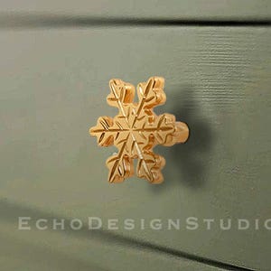 Op de afbeelding: Een goudkleurige kastknop in de vorm van een sneeuwvlok. De knop is gemonteerd op een groene kastdeur. De tekst "ECHO DESIGN STUDIO" staat in het wit onderaan de afbeelding.