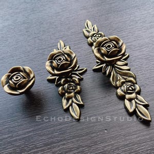 Puede incluir: Tres tiradores de cajón de color dorado antiguo con un diseño floral. Los tiradores presentan una sola rosa con hojas y rosas más pequeñas.