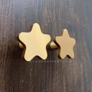 May include: Two gold-colored star-shaped cabinet knobs. The knobs are made of metal and have a brushed finish. The knobs are mounted on a dark brown wooden surface. The text "ECHODESIGNSTUDIO" is visible.