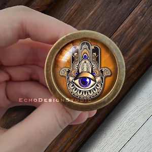 Peut inclure: Un bouton en métal doré avec un dessus rond en verre représentant un motif détaillé de main de hamsa avec un œil bleu au centre. La main de hamsa est entourée de motifs complexes et est placée sur un fond doré.