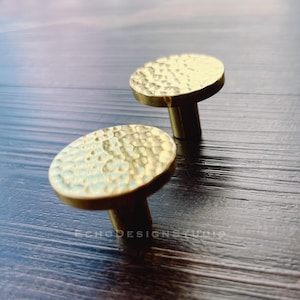 Op de afbeelding: Twee gouden ronde kastknoppen met hamermotief op een donker houten oppervlak.