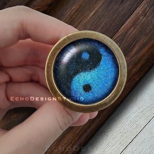 Puede incluir: Un pomo redondo de color latón con un símbolo yin yang azul y negro en la parte delantera. El símbolo está rodeado de una resina transparente y texturizada.