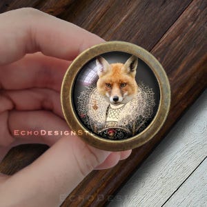 May include: A circular cabinet knob with an antique brass finish, showcasing a fox in formal attire. The fox has orange fur and wears a decorative collar. The text "ECHODESIGNST" is printed on the knob.