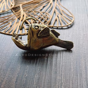 Puede incluir: Una figurita de pez pescador de color bronce con una boca grande y dientes afilados. La figurita está detallada con escamas y aletas texturizadas.