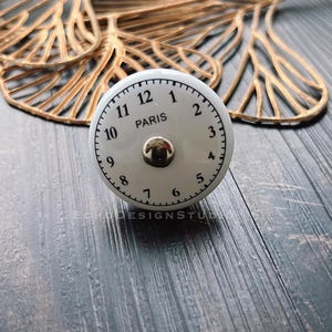 Puede incluir: Pomo de cerámica blanco con un diseño de esfera de reloj negro. La esfera del reloj tiene números negros y la palabra "PARIS" en el centro.