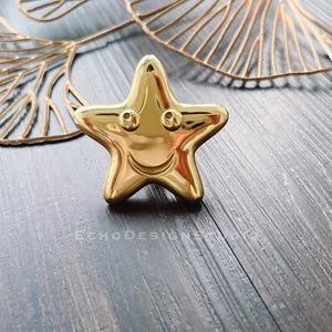 Puede incluir: Un objeto decorativo dorado brillante en forma de estrella con una cara sonriente. La estrella tiene dos pequeños agujeros para los ojos y una línea curva para la boca. El fondo incluye formas doradas en forma de hoja y una superficie de vetas de madera oscura.