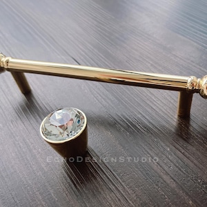 May include: Gold-toned cabinet hardware set featuring a long pull and a single knob. Both pieces have clear, faceted crystal accents and are set against a dark wood surface. The pull measures approximately 6 inches in length.