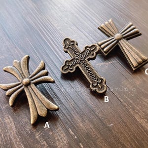 Op de afbeelding: Drie antieke, goudkleurige, kruisvormige metalen versieringen. De kruisen hebben verschillende ontwerpen en zijn gerangschikt op een houten oppervlak.