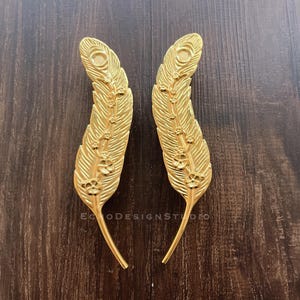 May include: Two gold-colored metal feather-shaped door pulls with intricate floral designs.