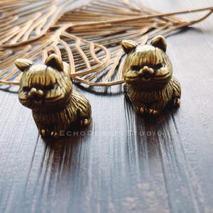 May include: Two small, detailed brass cat figurines. The cats are sitting upright with their ears perked up. The figurines have a textured, aged appearance. The background includes a gold-colored leaf and a dark wooden surface.