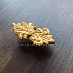 Puede incluir: Pomo de armario de metal dorado con un diseño floral decorativo y ornamentado.