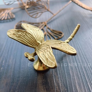 May include: A gold-toned dragonfly-shaped cabinet knob with intricate details. The knob is mounted on a round base.