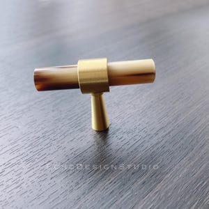 Könnte beinhalten: Ein Messing- und brauner Schrankknopf mit T-förmigem Design. Der Knopf hat eine glatte, polierte Oberfläche.