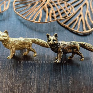 May include: Two brass fox figurines, one gold and one bronze, are displayed on a dark wood surface. The foxes are detailed with realistic features and are positioned side-by-side. Decorative brass leaf-shaped elements are visible in the background.
