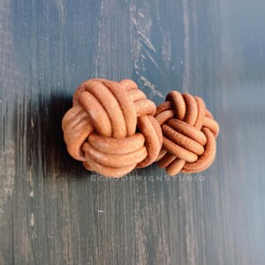 May include: Two brown leather knot-shaped cufflinks. The cufflinks are made of braided leather and are a light brown color. The text "ECHODESIGNSTUDIO" is visible in the image.