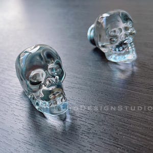 May include: Two clear glass skull bottle stoppers. The skulls are detailed with eye sockets, a nose, and a jawline.