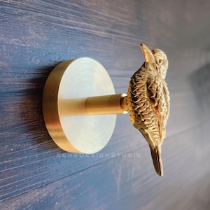 May include: A gold-colored wall hook featuring a detailed bird design. The hook has a circular base and a cylindrical arm, with the bird perched at the end. The hook is mounted on a dark blue wooden surface. The text "ECHODESIGNSTUDIO" is visible.