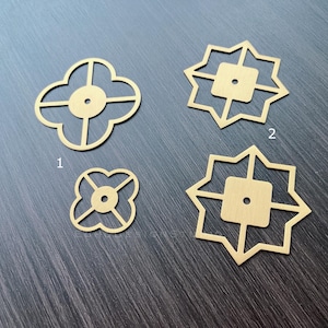 May include: Four gold metal decorative embellishments with geometric designs. Two of the embellishments are shaped like a four-leaf clover, and two are shaped like a star with eight points.