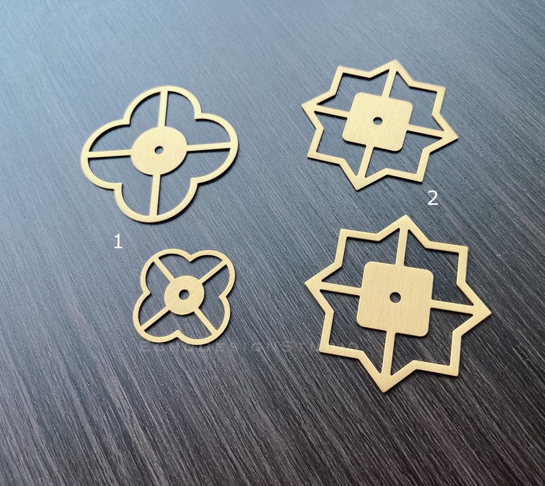 Puede incluir: Cuatro adornos decorativos de metal dorado con dise&ntilde;os geom&eacute;tricos. Dos de los adornos tienen forma de tr&eacute;bol de cuatro hojas, y dos tienen forma de estrella de ocho puntas.