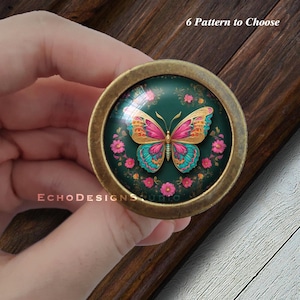 Puede incluir: Un pomo redondo de color latón con un inserto de vidrio que presenta una mariposa colorida con alas rosadas, azules y amarillas sobre un fondo verde con flores rosas. El texto "6 Pattern to Choose" es visible sobre el pomo.