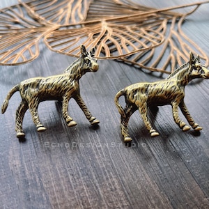 May include: Two bronze-colored donkey figurines with textured details. The donkeys are posed in a walking stance, with their legs spread. The background includes a decorative gold-colored leaf and a dark wooden surface.