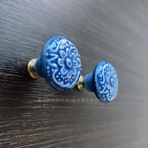 Könnte beinhalten: Zwei blaue Keramik-Schrankknöpfe mit einem weißen Blumenmuster. Die Knöpfe haben eine Messingbasis.