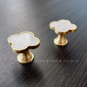 Könnte beinhalten: Zwei goldfarbene Schrankknöpfe mit einem weißen, perlmuttartigen Design aus vier Blättern.