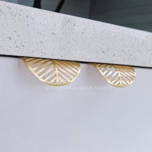 May include: Two gold-coloured leaf-shaped cabinet pulls are mounted on a white cabinet. The pulls have a cut-out design that resembles the veins of a leaf. The cabinet is under a white countertop.