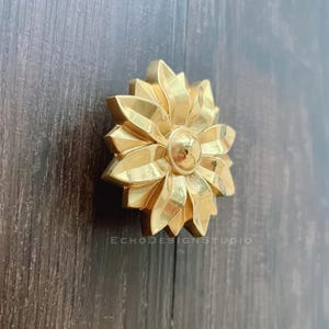 Op de afbeelding: Een gouden metalen bloemvormige kastknop met een verhoogd midden. De knop is gemonteerd op een donker houten oppervlak.