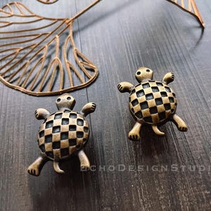 Könnte beinhalten: Zwei goldfarbene, schildkrötenförmige Schubladenknöpfe mit einem karierten Muster. Die Knöpfe befinden sich auf einer dunklen Holzoberfläche.