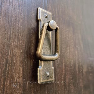 Puede incluir: Tirador de armario de latón antiguo con asa de anillo. El tirador tiene una base rectangular con detalles decorativos.