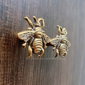 May include: Two brass bee-shaped cabinet pulls mounted on a dark wood surface. The bee pulls are detailed with wings, legs, and body segments. The pulls are in a vintage style and add a decorative touch to furniture.