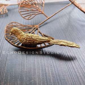 Pode incluir: Uma figurinha de pássaro de metal dourado pousada em uma escultura de folha de metal dourado. O pássaro tem uma cauda longa e está voltado para a direita. A escultura de folha é feita de metal fino e tem um design delicado.