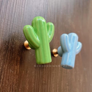 Può includere: Due pomelli per porte in ceramica a forma di cactus. Uno è verde e l'altro è blu.