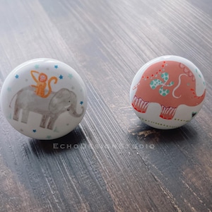 Pode incluir: Dois puxadores de cerâmica branca. Um puxador apresenta um elefante cinzento com um macaco laranja nas costas, rodeado por estrelas azuis. O outro puxador tem um design de elefante rosa com detalhes em turquesa.