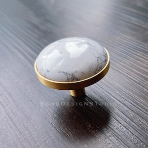 Op de afbeelding: Een ronde, witte marmeren kastknop met een gouden metalen basis. De knop heeft een wit en grijs marmerpatroon.