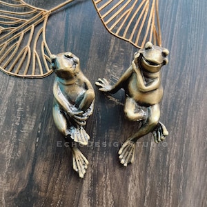 May include: Two gold frog figurines with a smiling expression. The frogs are sitting on a dark wood surface.