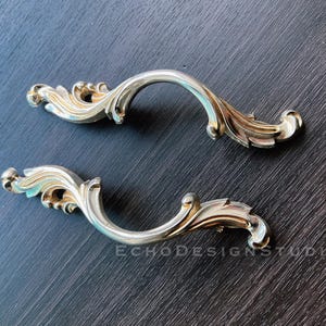 May include: Two ornate gold-toned cabinet pulls with a decorative scroll design. The pulls are made of metal and have a smooth, polished finish.