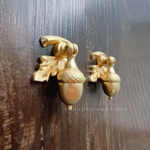 Puede incluir: Dos pomos de armario en forma de bellota dorados con detalles de hojas. Los pomos están montados en una superficie de madera oscura.
