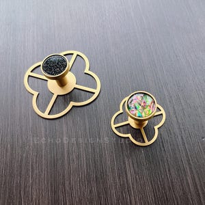 Puede incluir: Dos pomos de armario dorados con un dise&ntilde;o de tr&eacute;bol de cuatro hojas. Un pomo tiene un centro negro moteado y el otro tiene un centro iridiscente de colores.