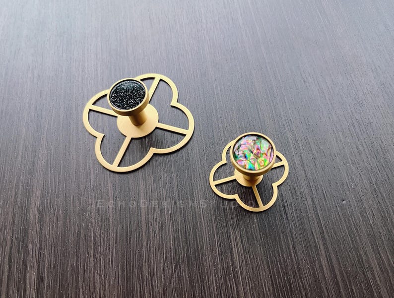 Puede incluir: Dos pomos de armario dorados con un dise&ntilde;o de tr&eacute;bol de cuatro hojas. Un pomo tiene un centro negro moteado y el otro tiene un centro iridiscente de colores.