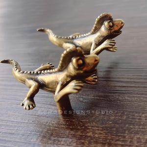 Puede incluir: Dos tiradores de armario con forma de lagarto de color bronce. Los lagartos tienen piel texturizada y extremidades extendidas. Están montados sobre una superficie de madera oscura, listos para su instalación.