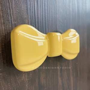 May include: A glossy, yellow bow-shaped decorative object. The bow has a rounded, three-dimensional design with a central knot and two loops. The text "ECHODESIGNSTUDIO" is printed on the bottom of the bow.