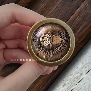 May include: A round, brass-coloured pendant with a glass dome. Inside the dome is a steampunk-style owl with gears and wings. The owl's eyes are made of small, round, metal gears.