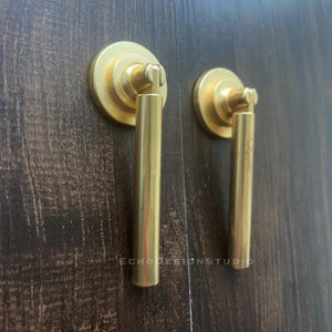 Puede incluir: Dos tiradores dorados para armario con base circular y asa cilíndrica. Montados sobre una superficie de madera marrón oscuro. El texto "ECHODESIGNSTUDIO" es visible en la madera.