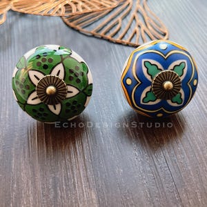 May include: Two ceramic knobs with intricate floral designs. One knob is green and white with a gold center, the other is blue and white with a gold center.