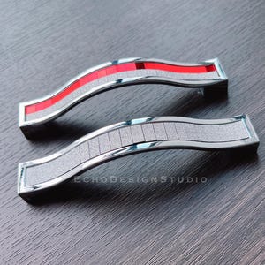 Puede incluir: Dos tiradores de armario plateados con un diseño ondulado. Uno de los tiradores presenta un centro rojo segmentado, mientras que el otro tiene un centro gris segmentado. Los tiradores tienen un marco rectangular y están sobre una superficie de madera oscura.
