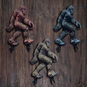 Puede incluir: Tres perchas de pared con forma de Bigfoot en varios colores: marrón rojizo, gris oscuro y beige. Cada percha es una escultura detallada de la criatura legendaria, montada en un gancho negro. Las perchas están montadas sobre una superficie de madera.